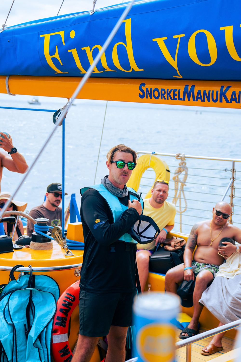 Man in sunglasses holds snorkel gear on a yellow Snorkel Manu Kai catamaran with passengers relaxing in background.