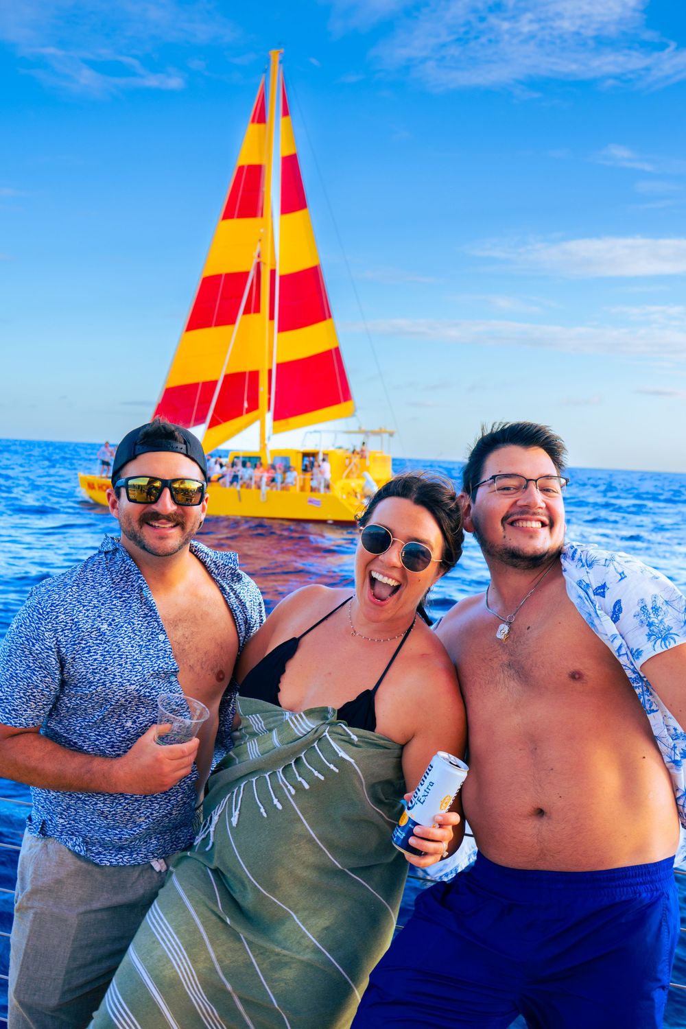 Three friends laughing and holding drinks on a catamaran with a red and yellow sailboat behind them.