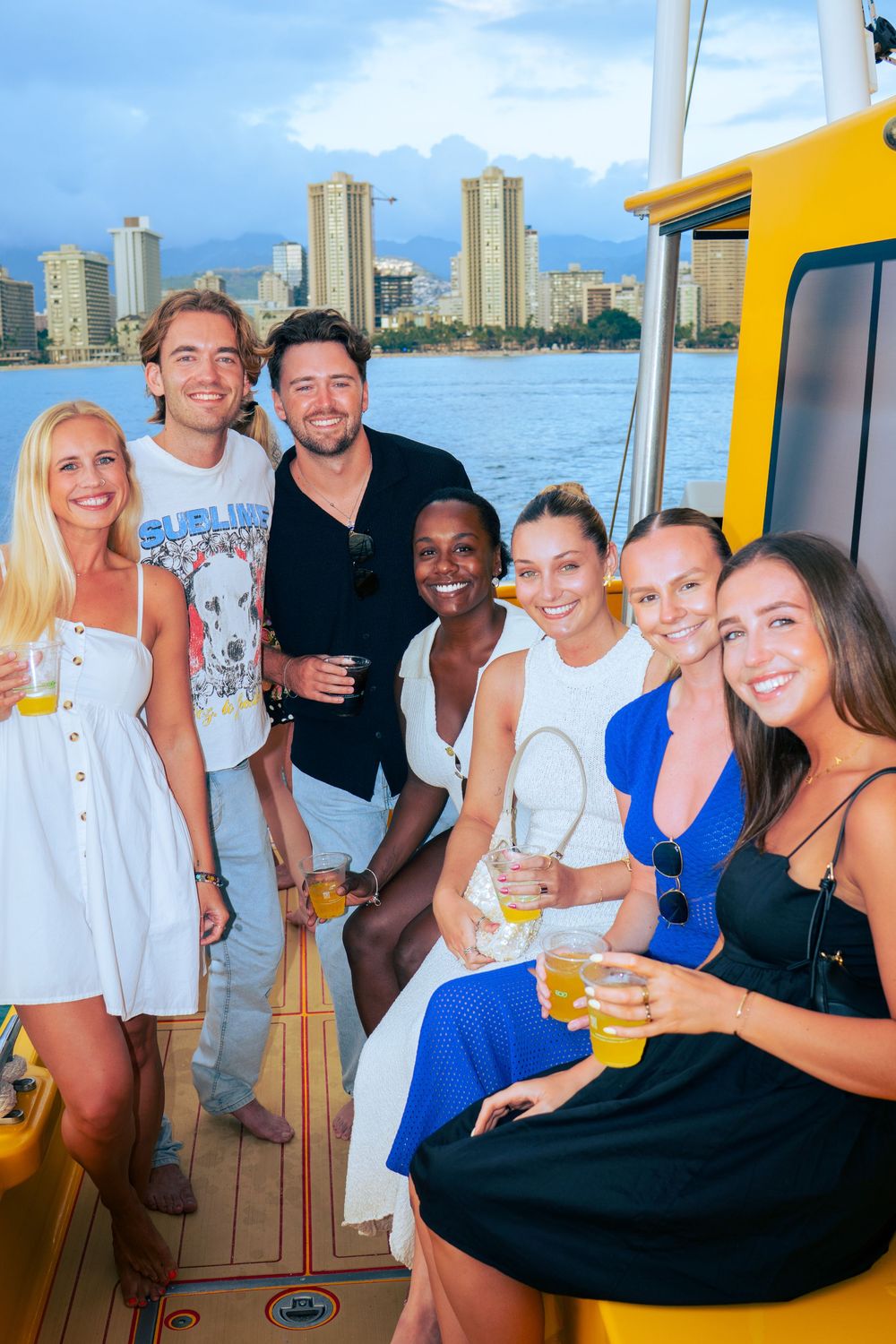 Group of seven friends smiling with drinks on a yellow catamaran at dusk with Waikiki skyline.