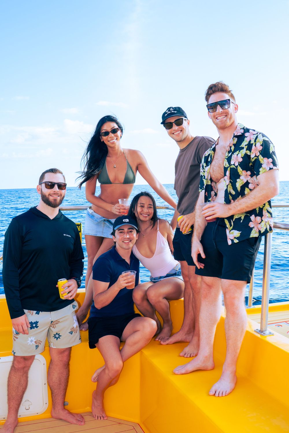 Six friends smiling and holding drinks on a yellow catamaran deck on a sunny ocean day.