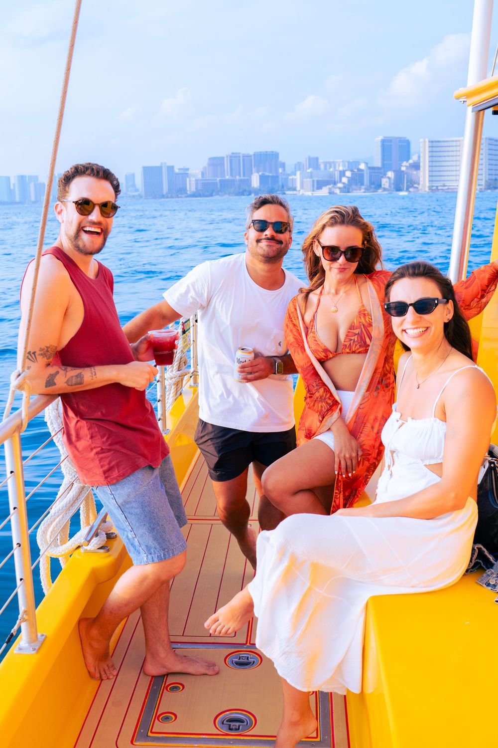 Group of four friends enjoying drinks on a yellow catamaran with a city skyline in the background.