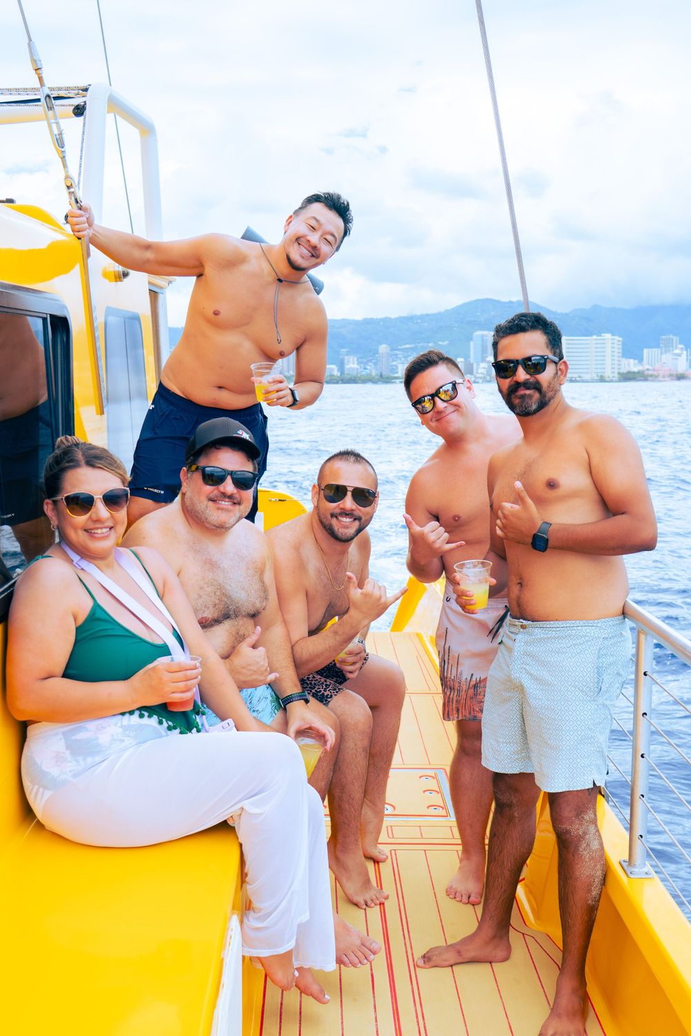 Group of six friends posing with drinks on a yellow catamaran with Honolulu skyline behind them.