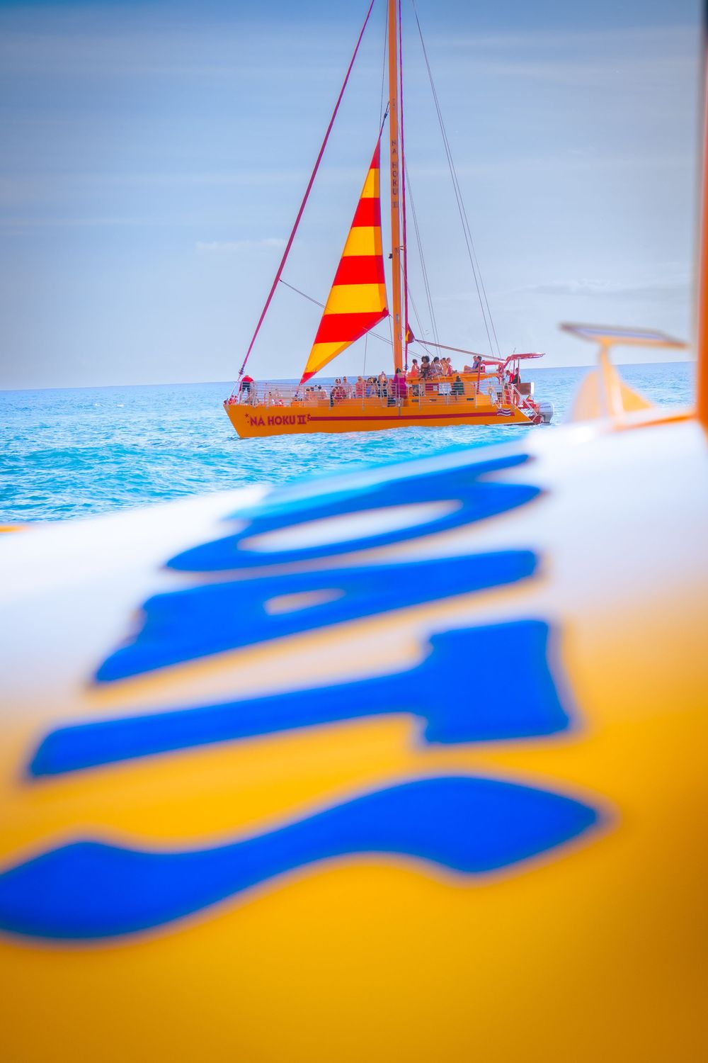 Colorful sailboat with orange and red striped sail on calm blue ocean seen from catamaran deck.