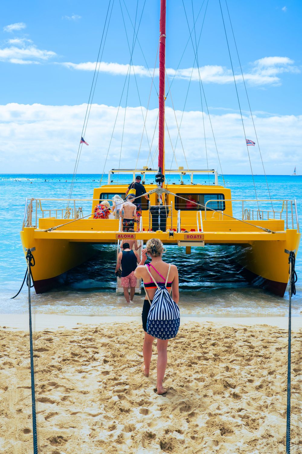 People boarding a bright yellow catamaran sailboat on a sandy Hawaiian beach with turquoise water.