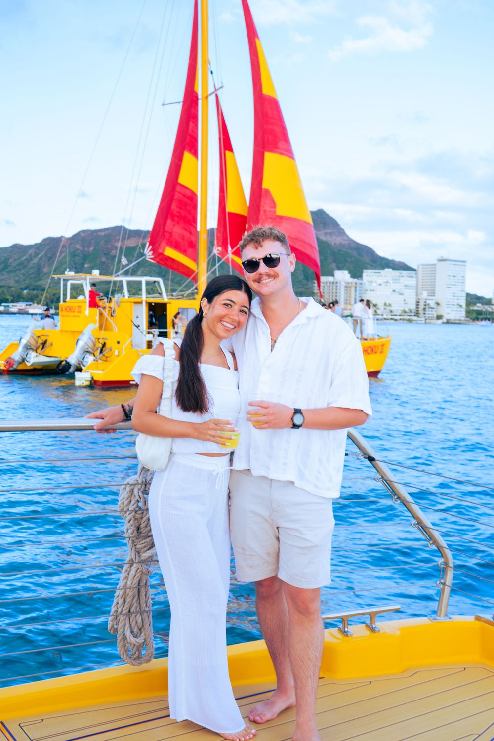 Couple in white outfits posing on a yellow catamaran sailboat with red and yellow sails in Hawaii.