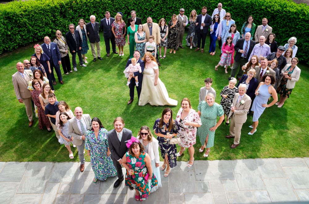 Aerial view of wedding guests forming heart shape on lawn around couple.