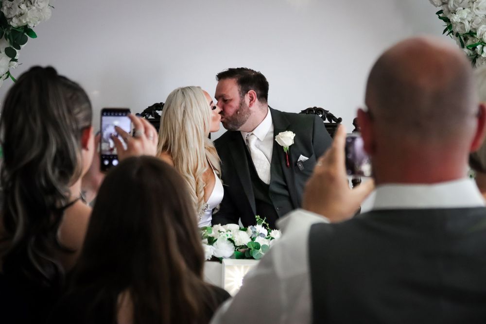 Wedding guests capture first kiss moment on smartphones during reception.