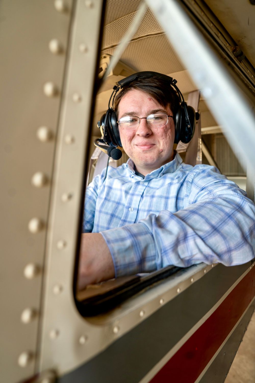 Young man wearing headphones and a plaid shirt smiling from inside a small aircraft window.