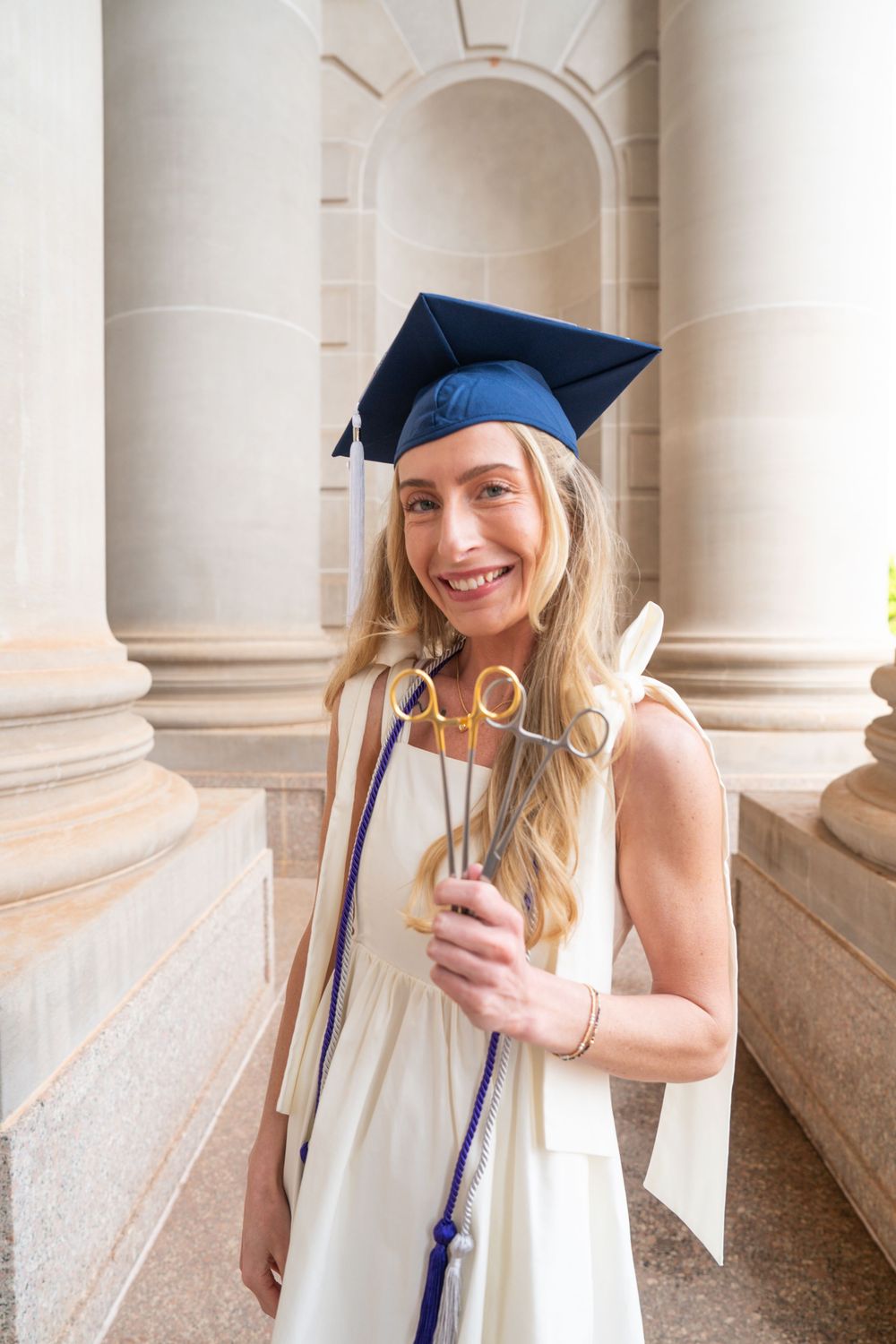 Smiling female graduate in navy cap and gown holding sparklers beside stone columns at graduation ceremony.