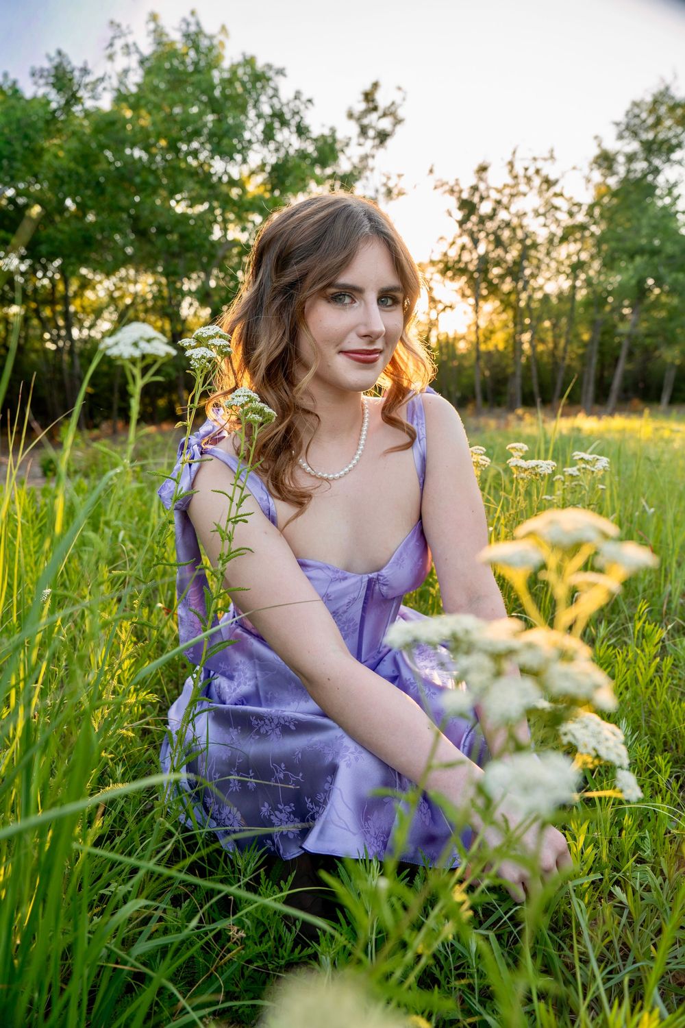 Young woman in lavender dress sitting in green meadow with wildflowers at golden hour sunset.