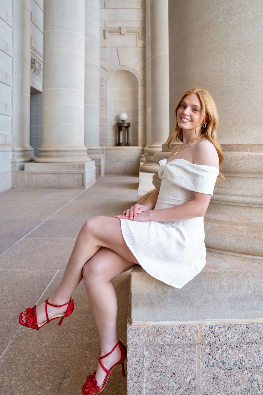 Young woman in white off-shoulder dress and red heels sitting on stone steps, smiling outdoors near columns.