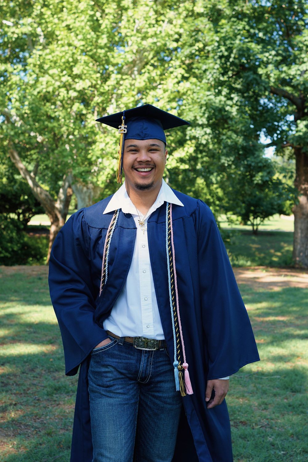 Graduate wearing blue cap and gown with honors cord standing outdoors on sunny day with trees in background.