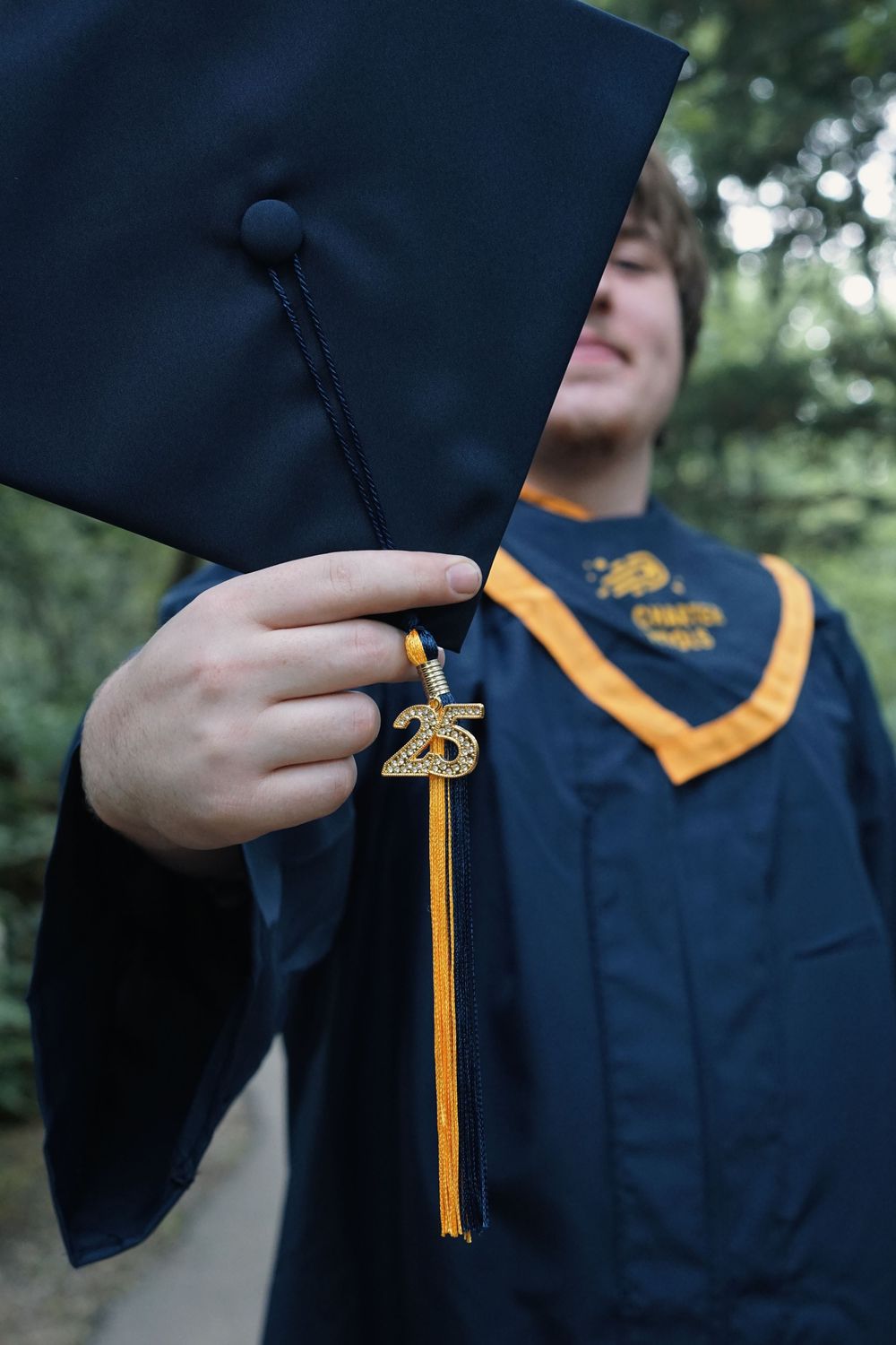 Graduate in blue cap and gown holds graduation tassel with gold honor cords against blurred outdoor background.