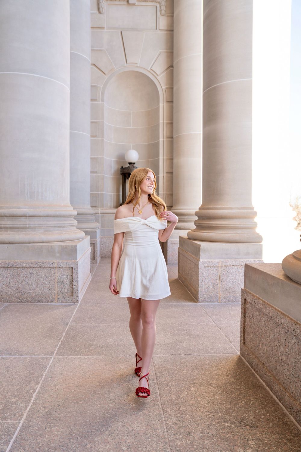 Young woman in white off-shoulder dress and red heels posing between grand stone columns of a classic building.