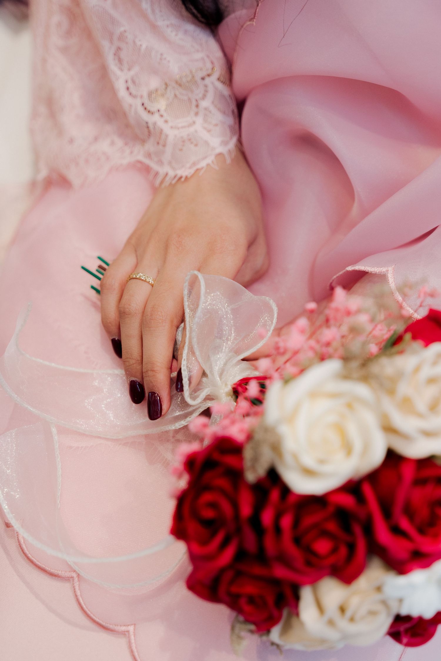 A bride in a pink lace dress holds a bouquet of red and white roses, showcasing a gold ring and dark manicured nails.