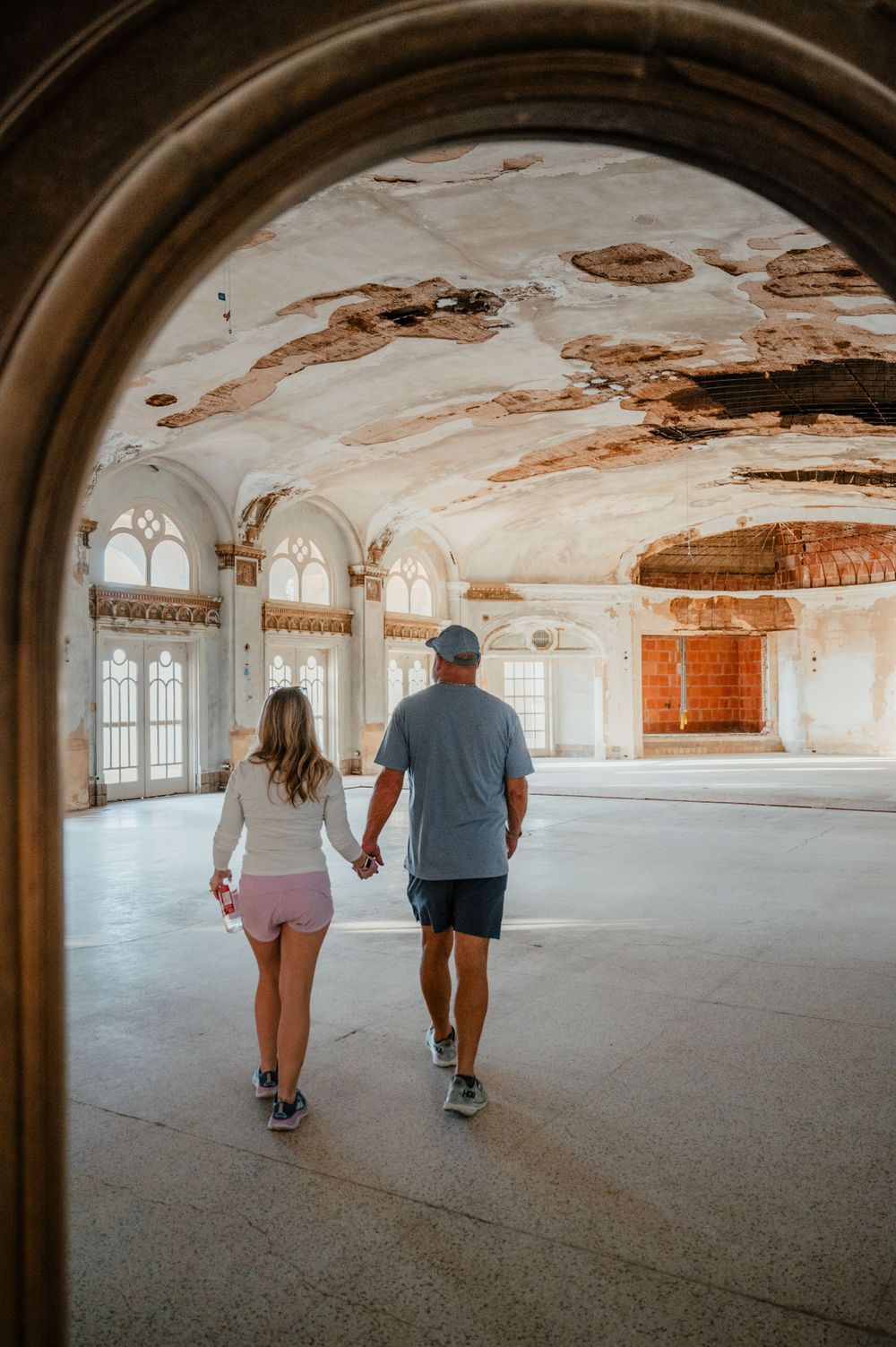 Two people exploring a grand empty room with ornate arched windows and deteriorating walls and ceiling.
