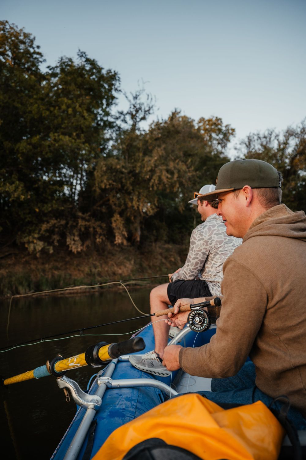 Two people sit together fishing from a boat on a lake with yellow gear and trees in background at sunset.