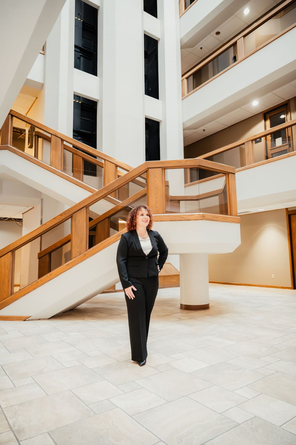 Professional in a dark business suit stands in modern office building lobby with wooden railings and marble floors.