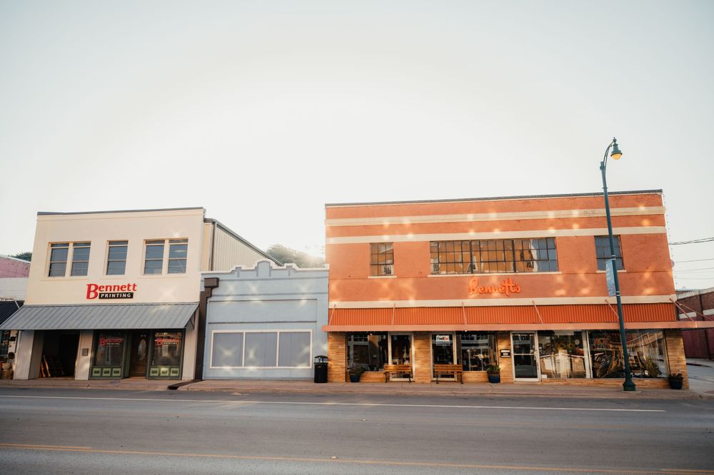 Small-town storefront buildings with vintage architecture and orange brick facade line a quiet street at dusk.