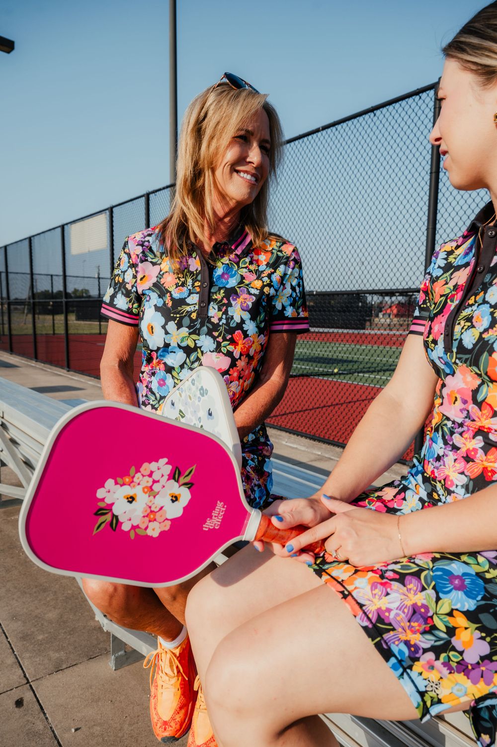 Two people in matching floral dresses sitting outdoors with pink pickleball paddle and tennis courts in background.