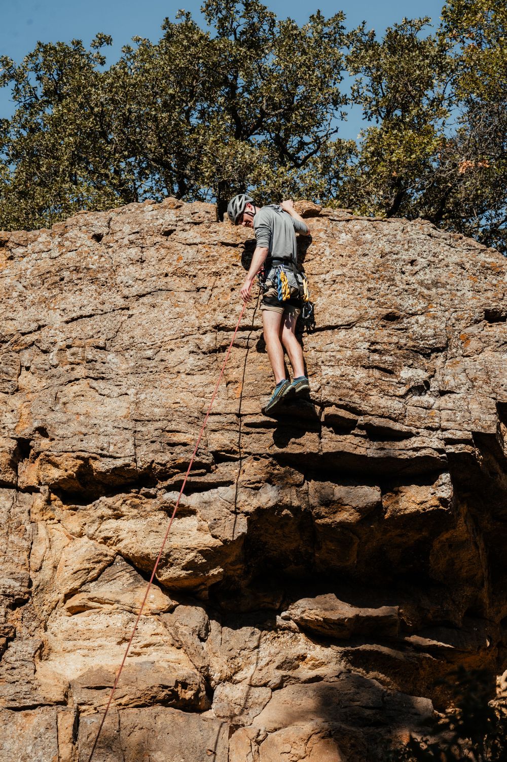 Rock climber scaling a steep rocky cliff face with climbing gear and ropes against a backdrop of green trees.