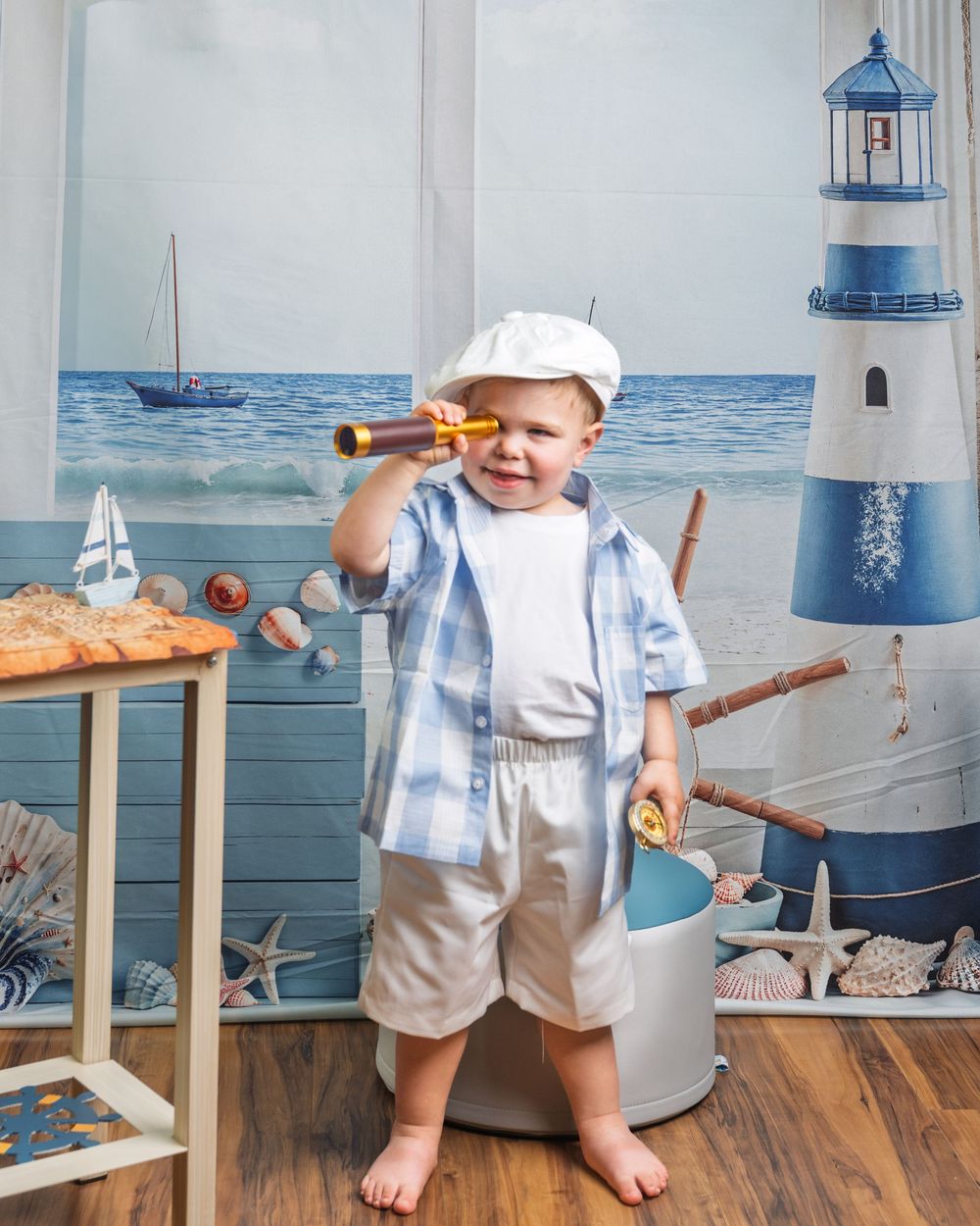 Toddler boy in sailor outfit holding telescope beside lighthouse decor with nautical beach backdrop.