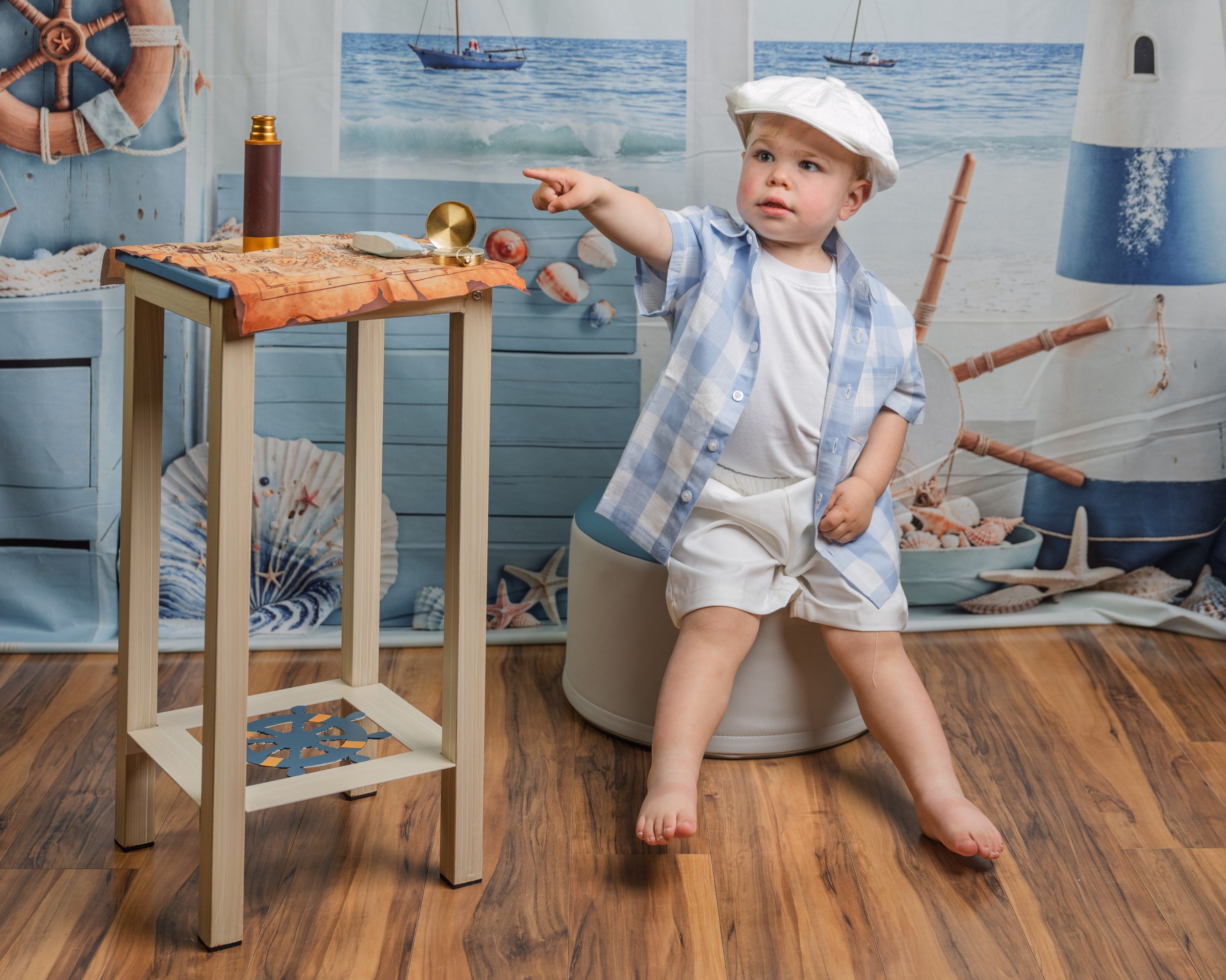 Toddler boy in blue shirt and white cap poses in nautical-themed studio with wooden stool and ocean decor.