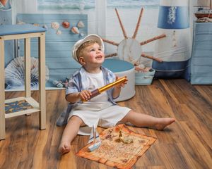 Toddler boy in sailor outfit holding telescope, sitting on floor with treasure map in nautical themed room.