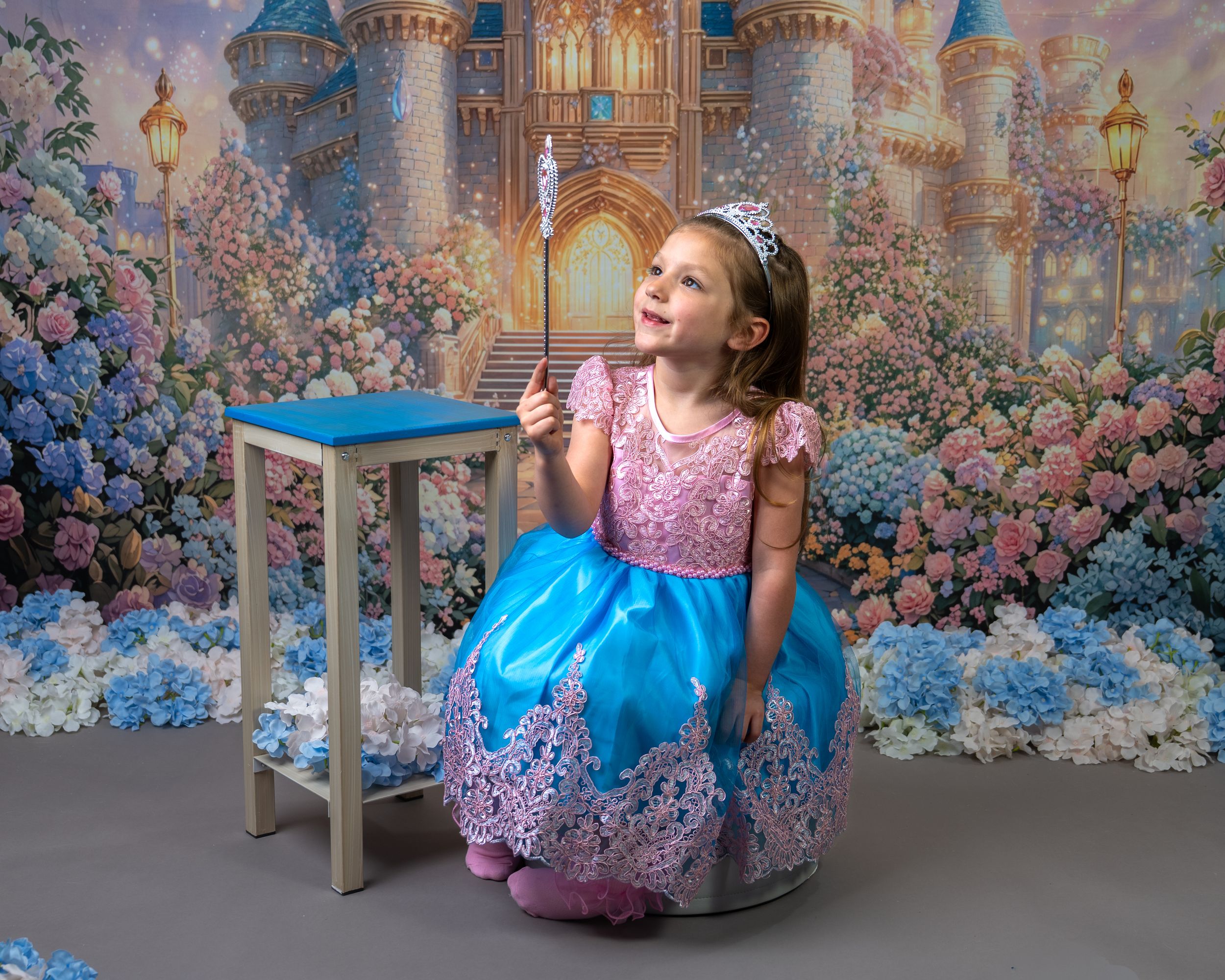 Young girl in blue and pink princess dress holding a wand, posing before a fairy tale castle backdrop.