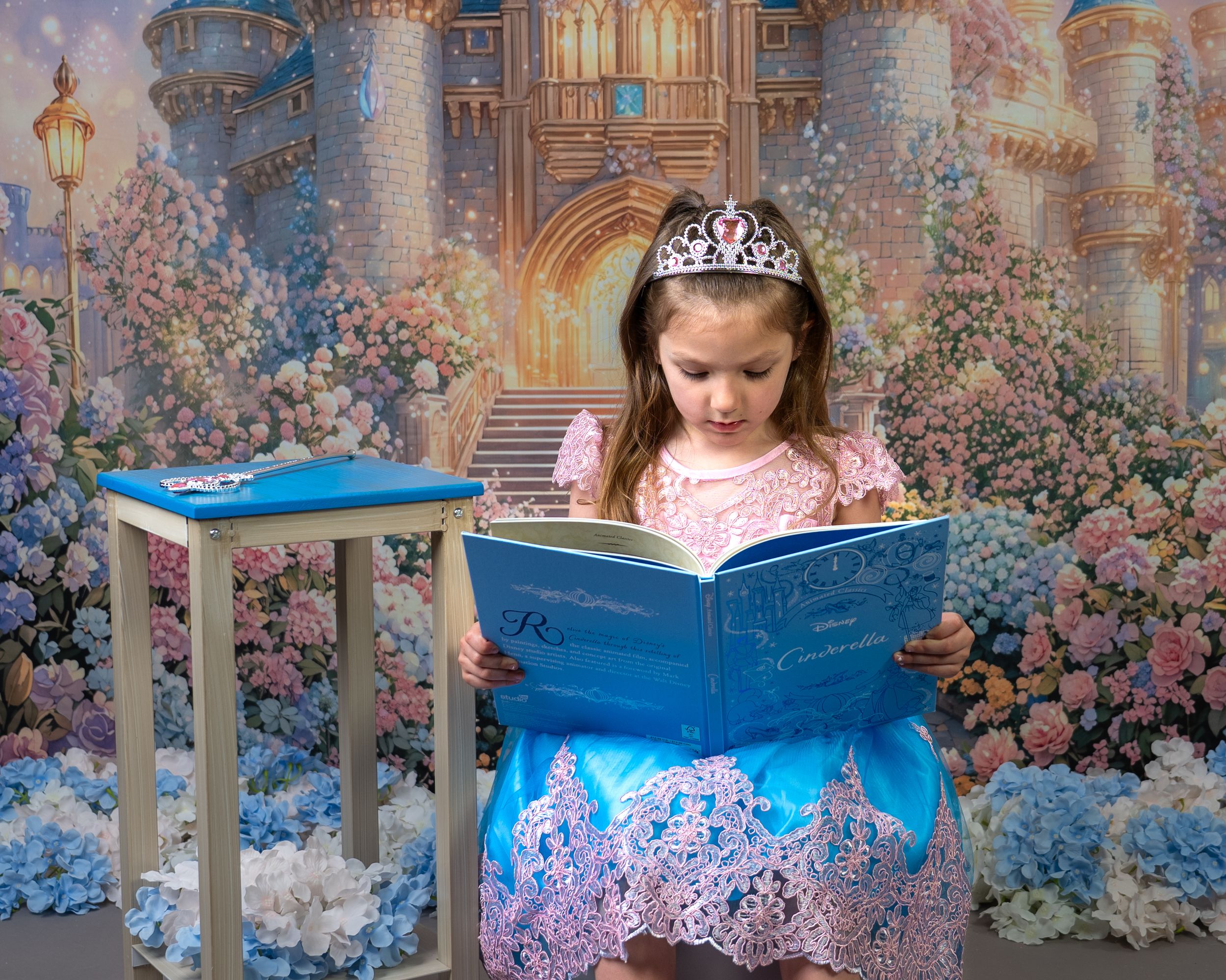 A young girl in a pink princess dress and tiara reads a blue storybook before a magical castle backdrop.