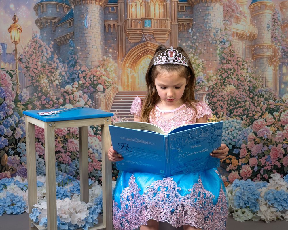 A young girl in a pink princess dress and tiara reads a blue storybook before a magical castle backdrop.