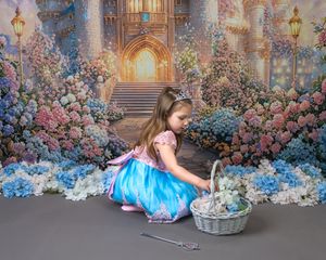 Young girl in blue princess dress kneeling by wicker basket before enchanted castle backdrop with colorful flowers.
