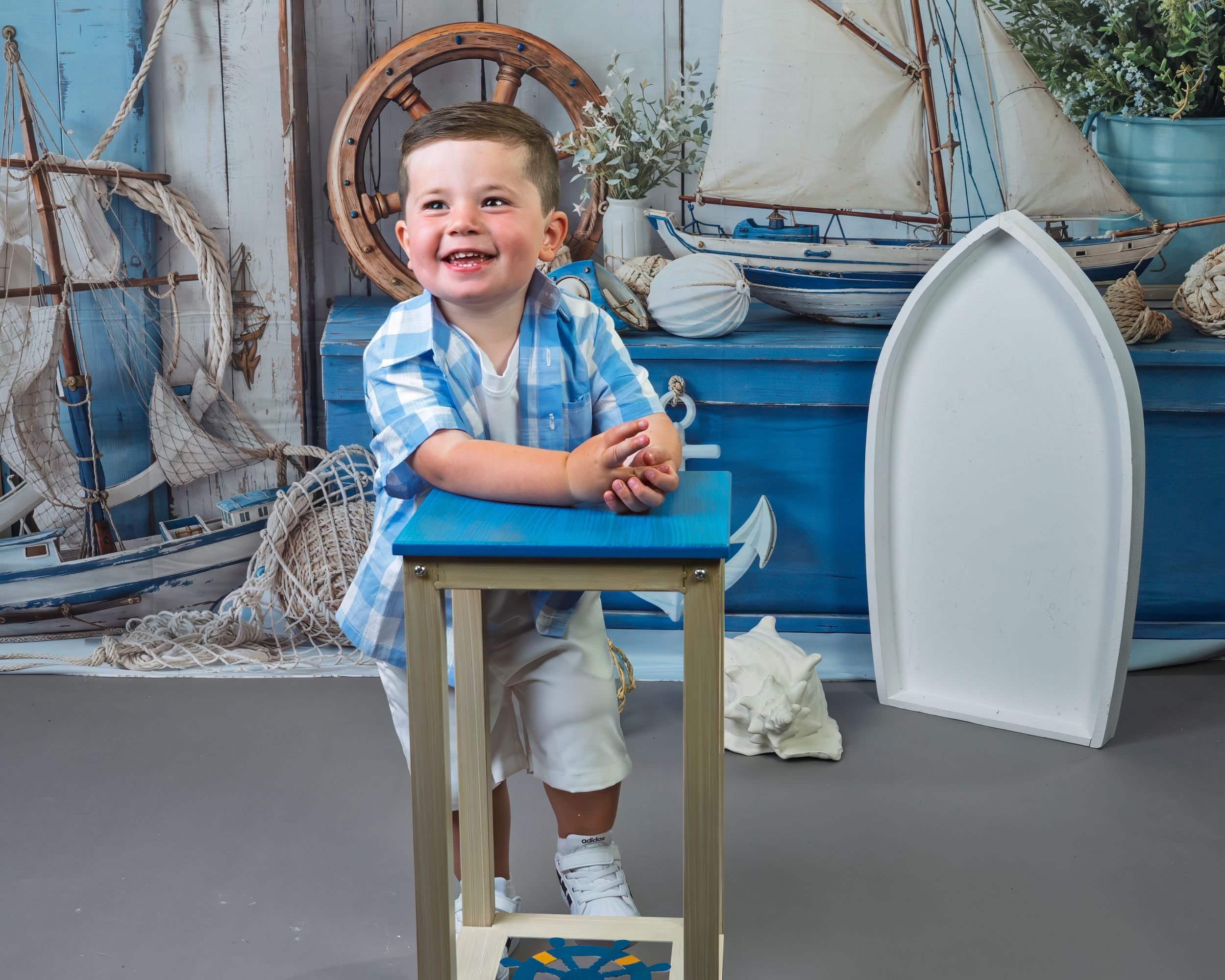 Smiling toddler boy in blue plaid shirt sits at small blue table with nautical themed backdrop featuring sailboats.