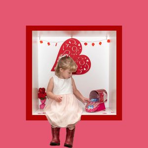 Young child in a pink dress stands against a pink background with Valentine's Day themed heart decorations.