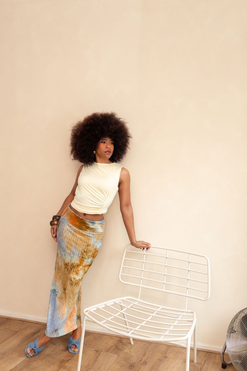 Woman with afro in white crop top and tie-dye maxi skirt standing beside white wire chair against beige wall.