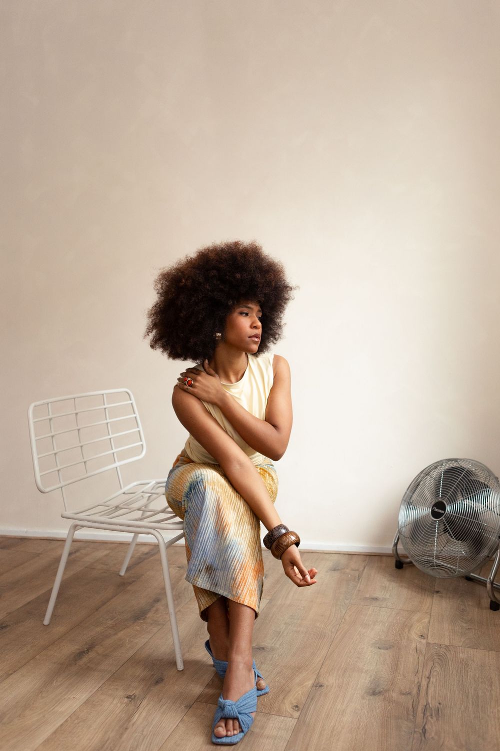 A woman with an afro sits on a white chair in a minimalist room, wearing a gold dress and blue sandals.