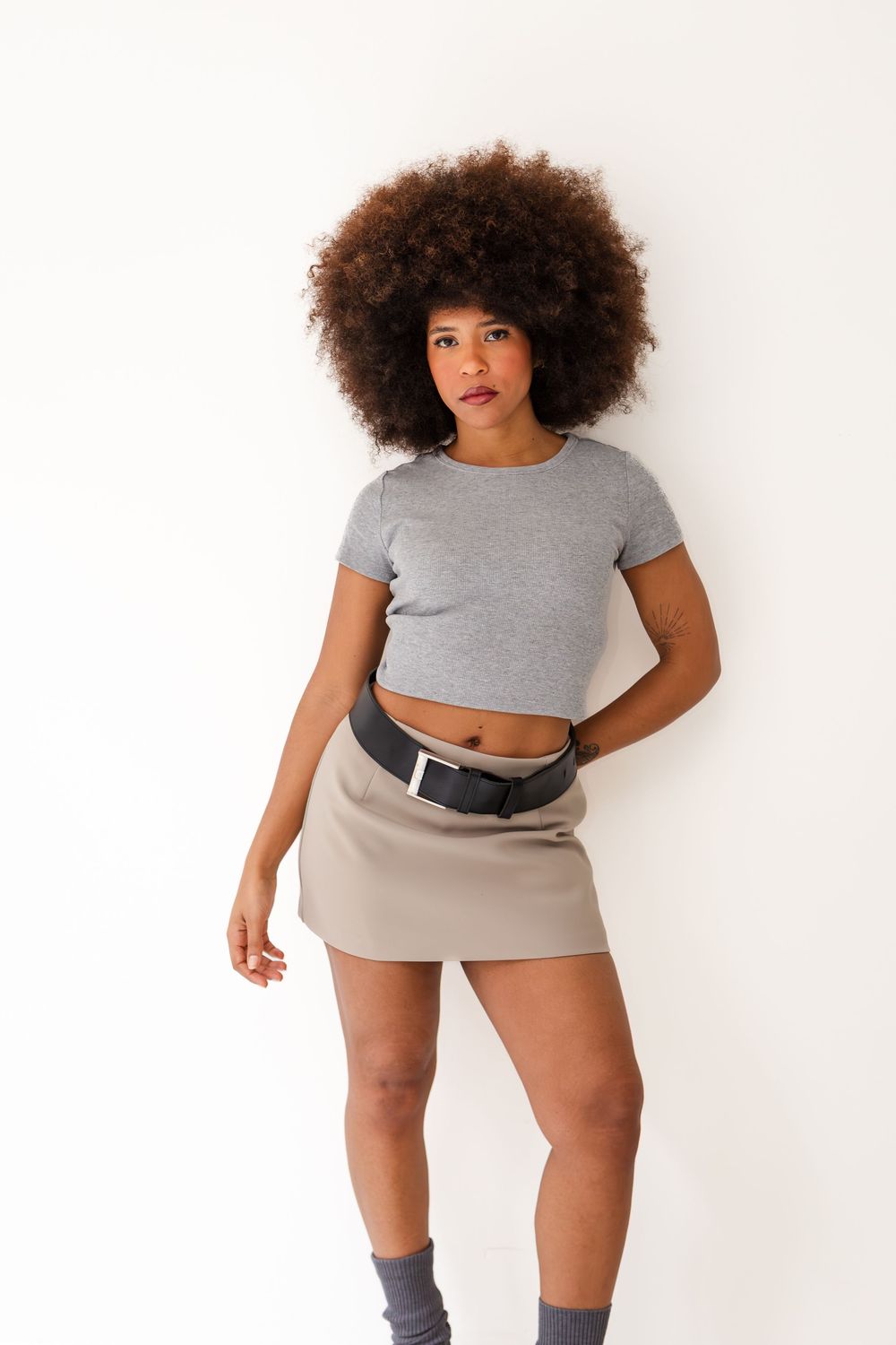 Woman with afro hair wearing gray crop top, beige mini skirt, and black belt bag against white background.