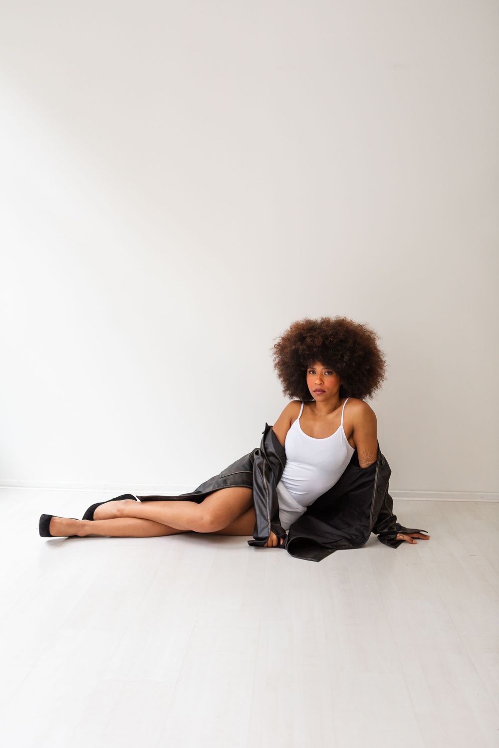 Young Black woman with afro hair sitting on floor wearing white bodysuit and black jacket in minimalist studio.