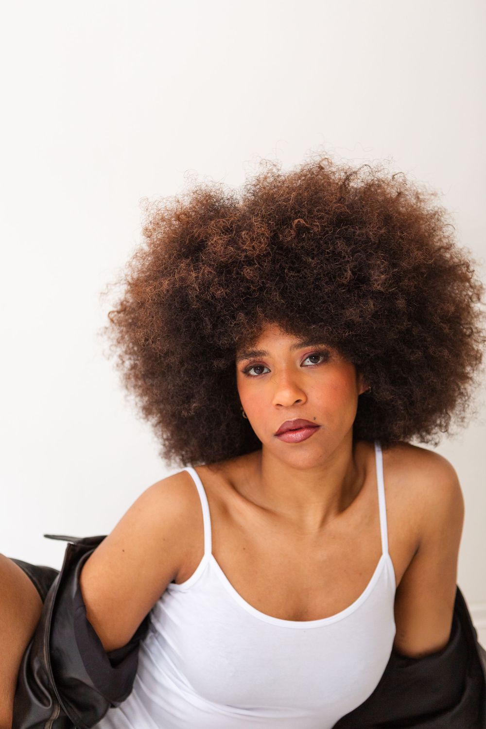 Young Black woman with large afro hairstyle wearing white tank top and black jacket, posing against white background.