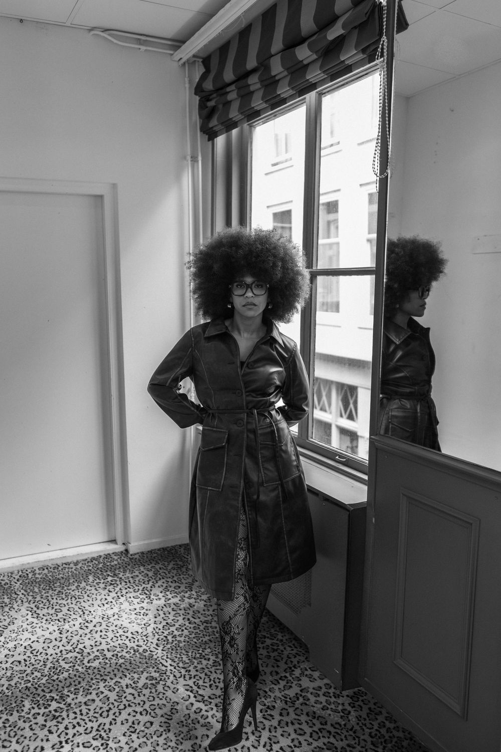 Black and white photo of a woman with an afro and glasses in a dark dress posing by a window with her reflection visible.