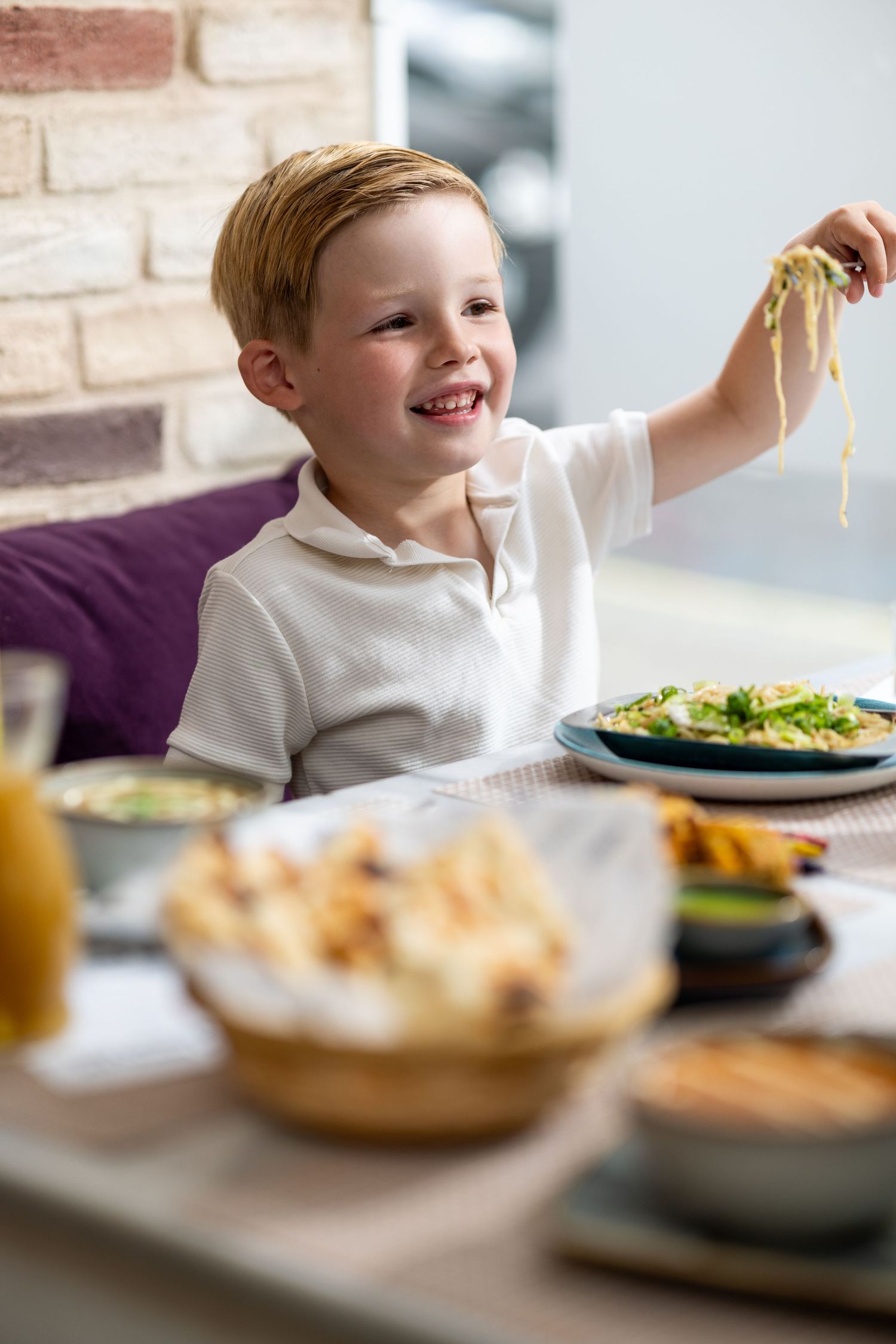 Young boy smiling and holding up pasta at a restaurant table, family dining lifestyle photography Southampton