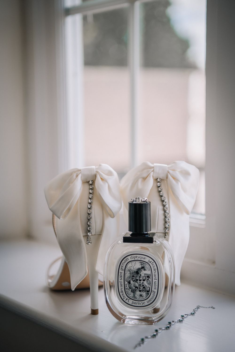Elegant bridal heels with white satin bows and crystal details beside a Diptyque perfume bottle on a windowsill.