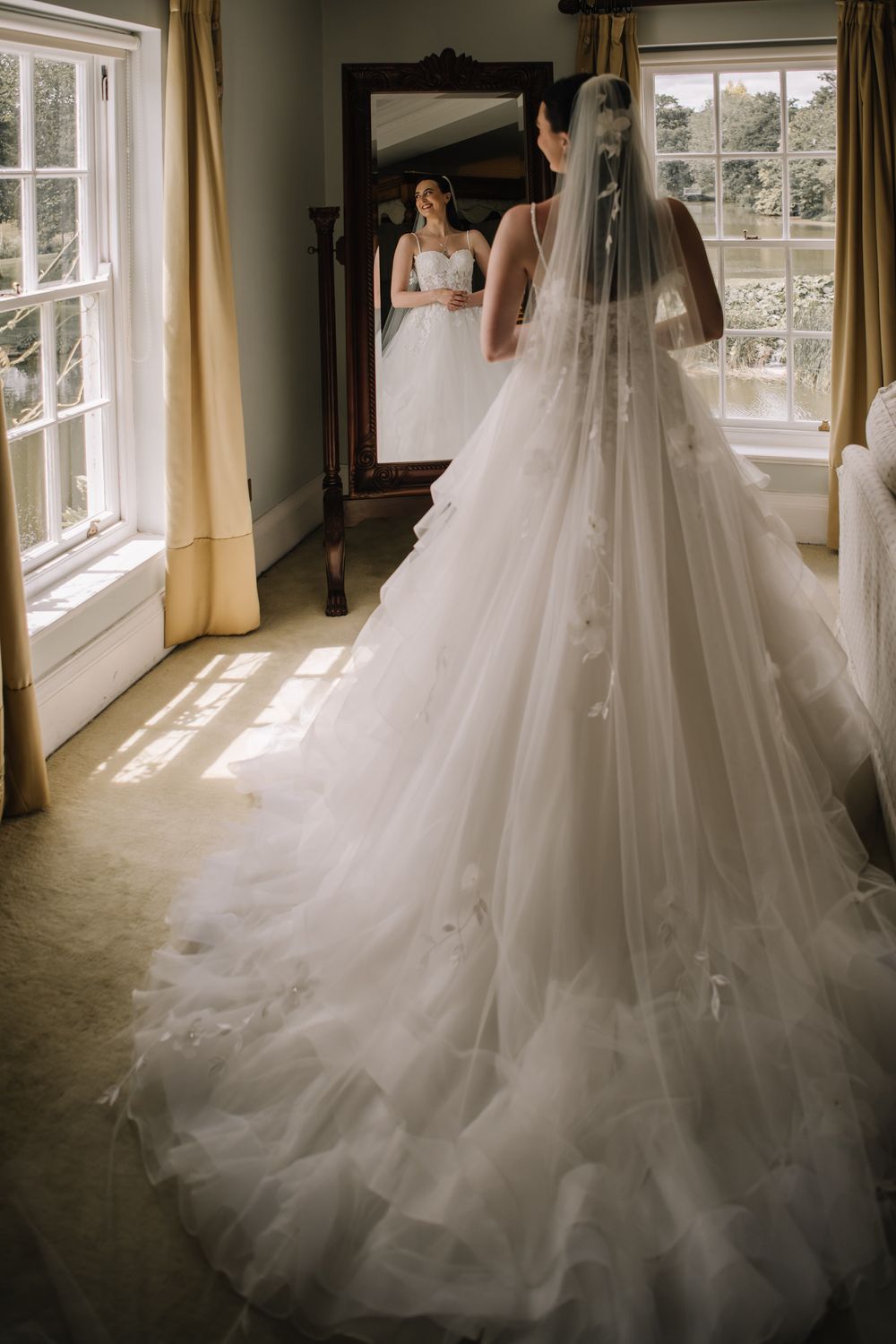 Bride in elegant white ballgown with long veil and floral train stands before a full-length mirror in a sunlit room.