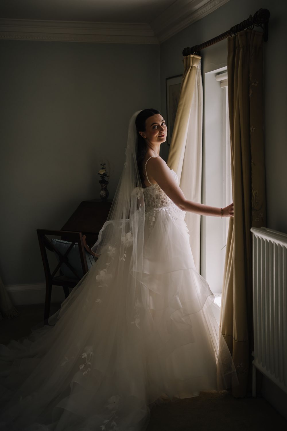 Elegant bride in layered tulle wedding dress and veil standing by a sunlit window in a softly lit room.