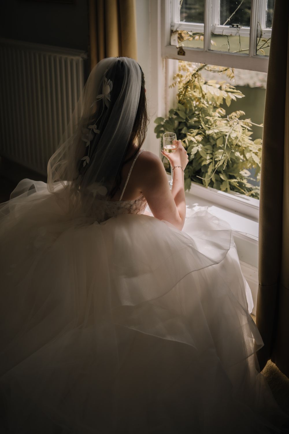 Bride in white gown and veil sits by window holding champagne glass, gazing at lush greenery outside.