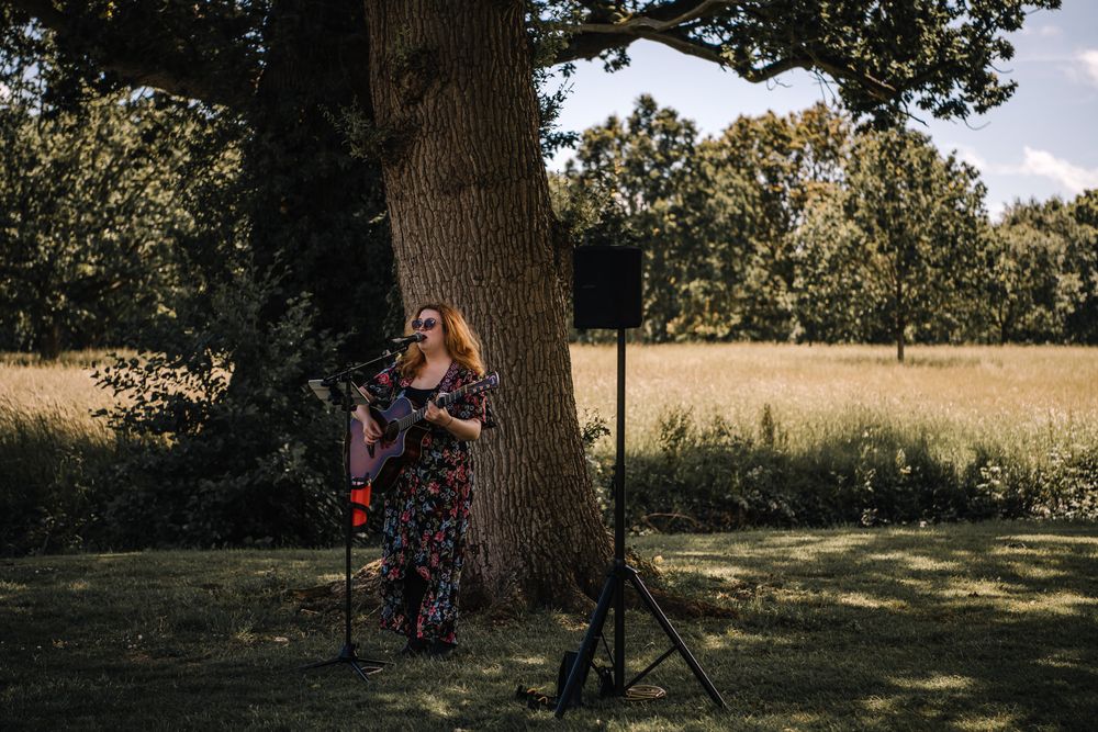 Female musician playing guitar outdoors by a large oak tree in a lush green field at a garden event.