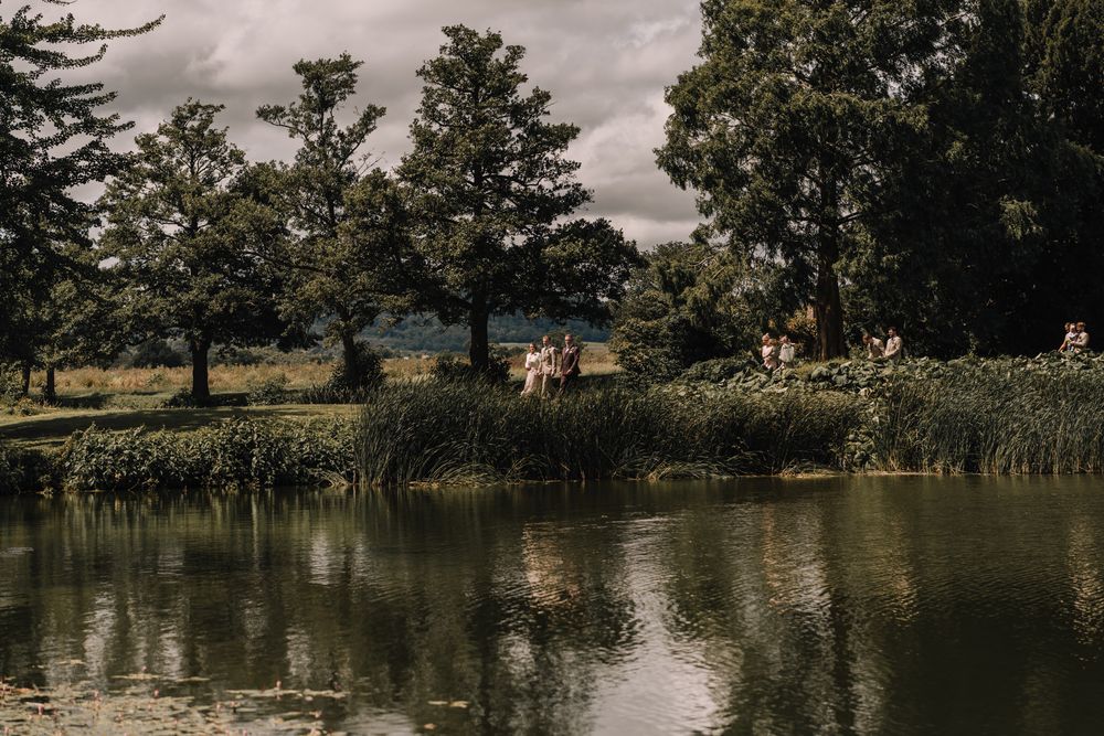 Serene countryside pond with lush green trees and guests gathered on the bank under cloudy skies.
