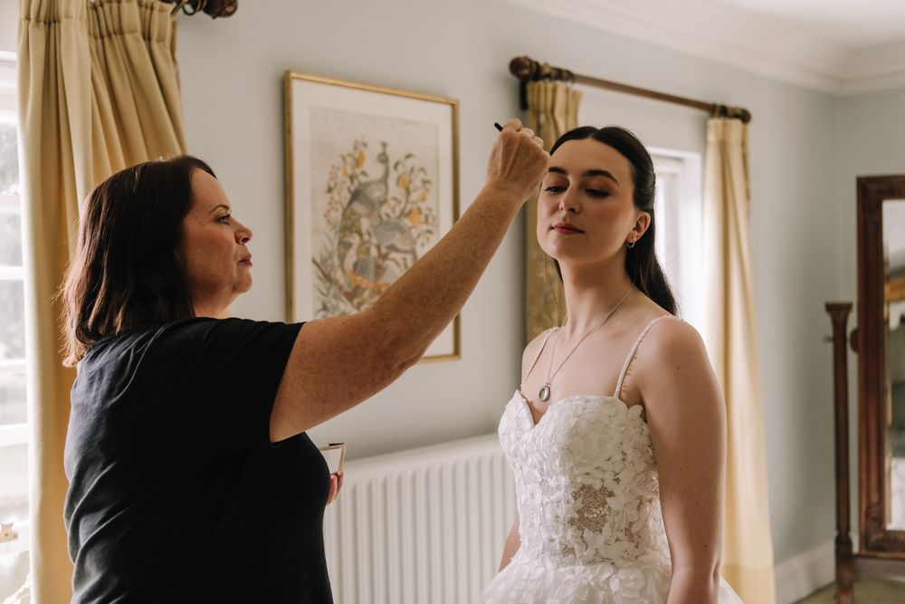 A makeup artist applies finishing touches to a bride in a white lace wedding dress before her ceremony.