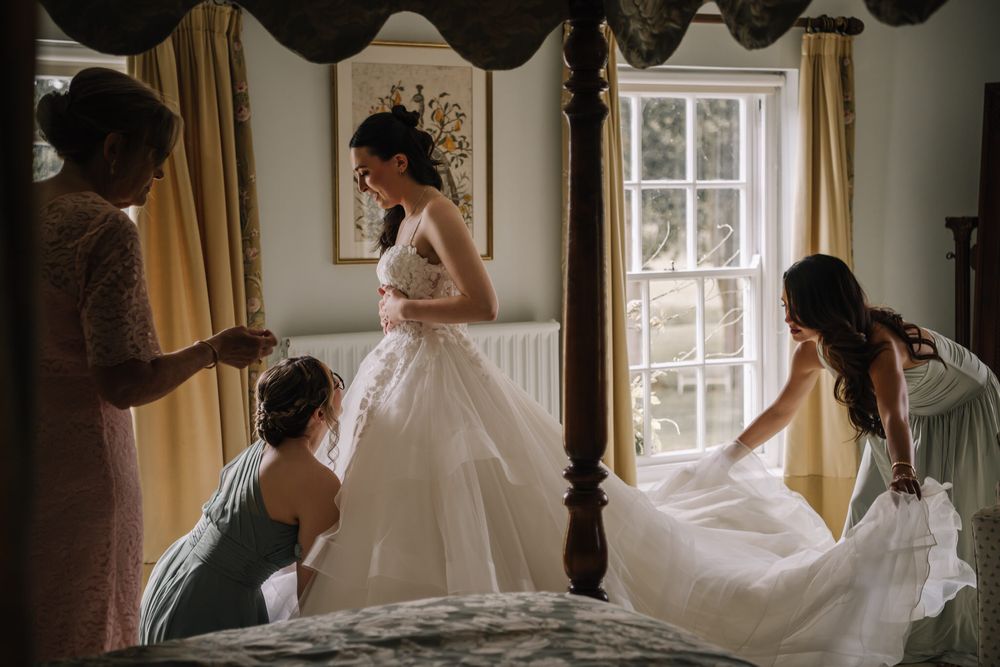 Bride in white ball gown being helped by bridesmaids in elegant bedroom with four-poster bed and natural window light.