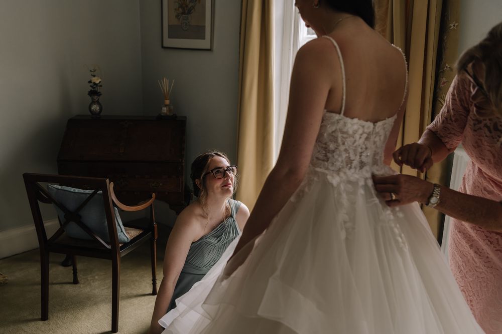Bride in white lace wedding gown being helped by two women near a window in a softly lit room.
